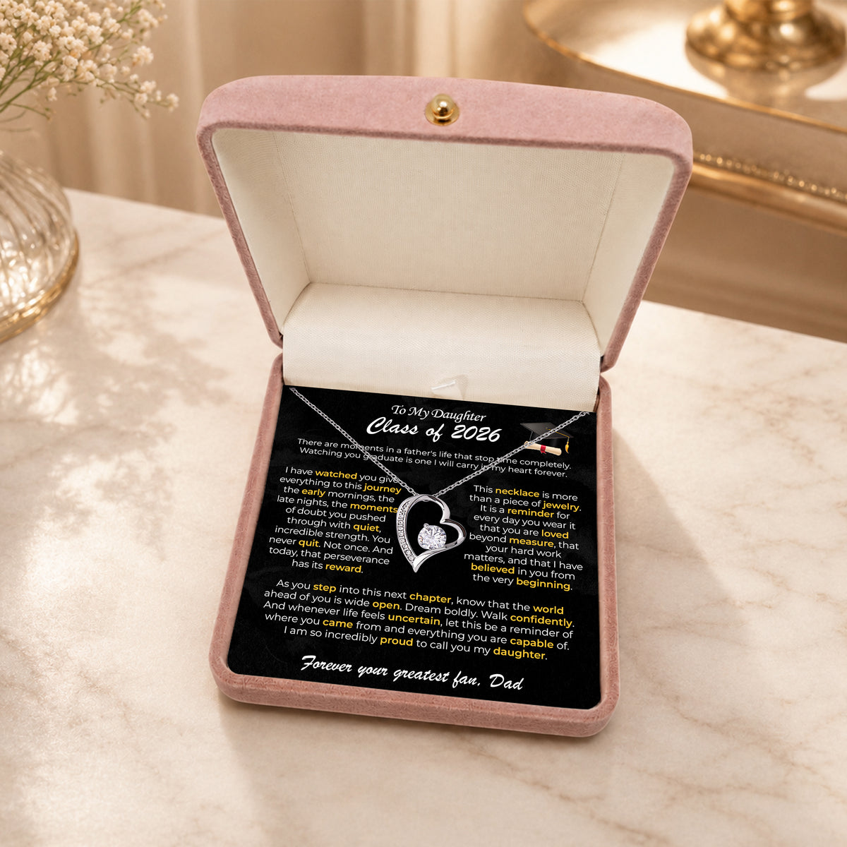 Gold heart pendant on a To My Daughter graduation card in a wooden box, in front of a pink bag with a scarf bow