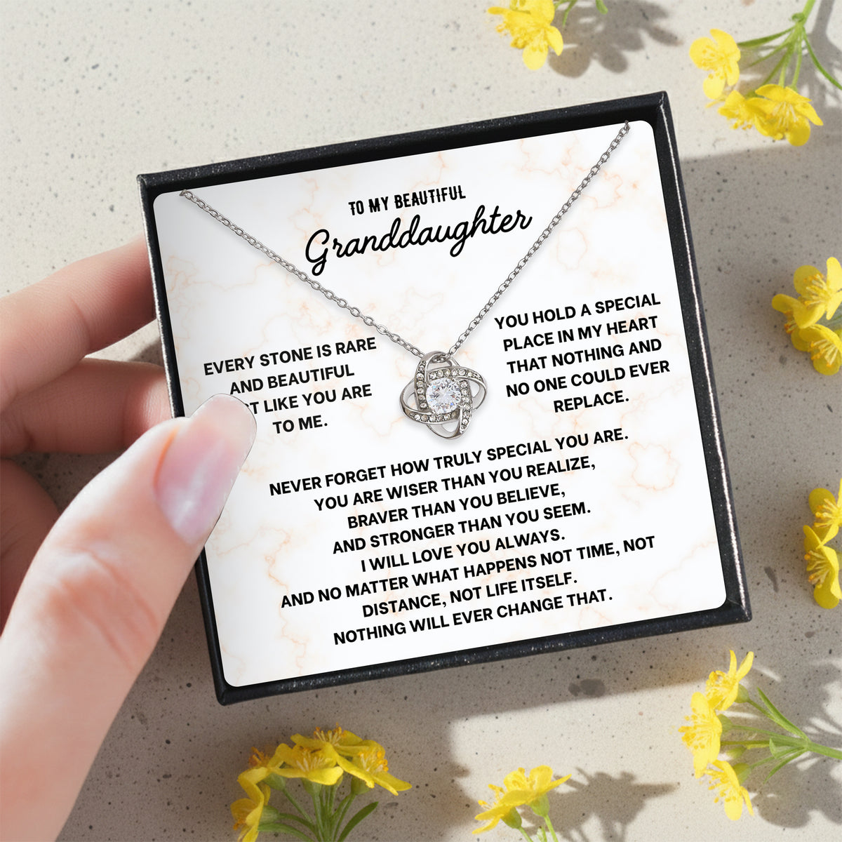 Hand holding a black gift box with a granddaughter necklace and message card, yellow flowers below